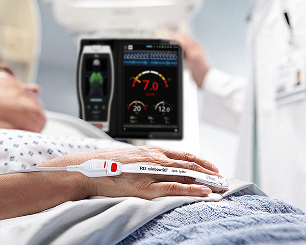 Patient im Krankenhausbett wird von einem Masimo-Sensor mit Root-Gerät überwacht und der Arzt im Hintergrund prüft die Daten auf dem Root-Gerät