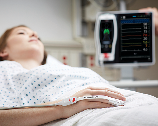 Schlafender Patient im Krankenhausbett, der einen Masimo-Fingersensor trägt, mit einem Root-Gerät im Hintergrund.