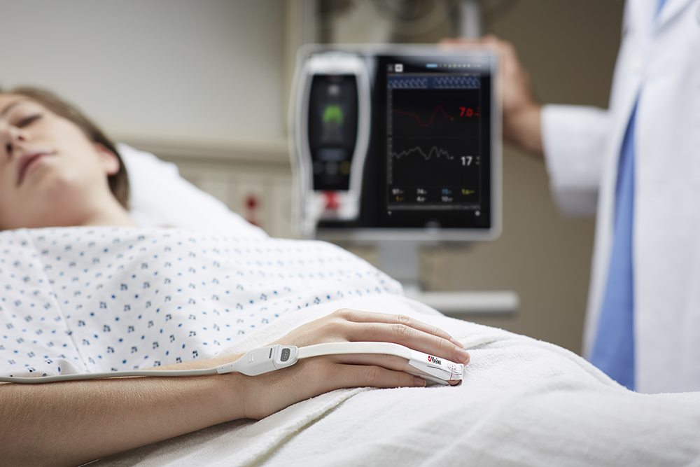 Schlafender Patient im Krankenhausbett mit Masimo-Monitor am Finger und einem Masimo-Root-Gerät im Hintergrund.